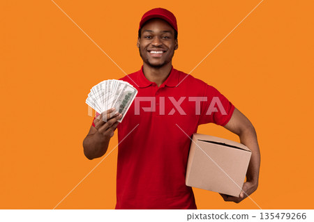 Cash On Delivery. Smiling african american male courier in red cap and uniform holding money and cardboard parcel box, carrying package, standing isolated over orange studio background 135479266