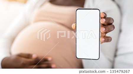 Pregnancy App. Black pregnant lady demonstrating smartphone with blank white screen, recommending new app for expecting women, demonstrating maternity shopping website, mockup image with copy space 135479267