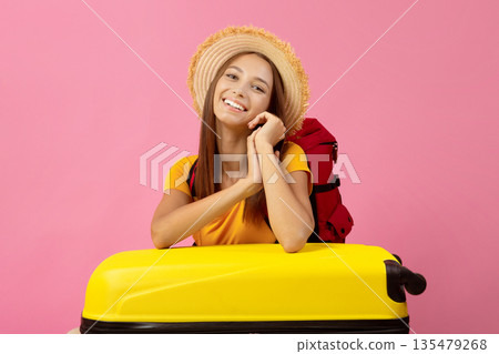 Happy young woman backpacker in summer hat leaning on big yellow suitcase and smiling at camera, pink studio background, copy space for advertisement. Cheerful female traveler going vacation 135479268