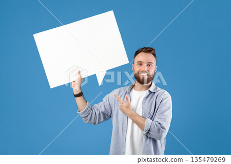 Happy young guy pointing at empty speech bubble with space for your ad or promo over blue studio background, mockup. Handsome Caucasian man with blank word cloud making announcement 135479269