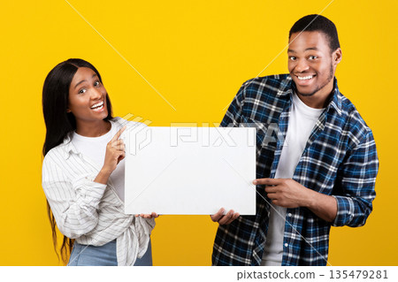 Interesting event, great offer and advice from couple. Cheerful millennial african american family in casual hold banner, pointing fingers to free space, isolated on yellow background, studio shot 135479281