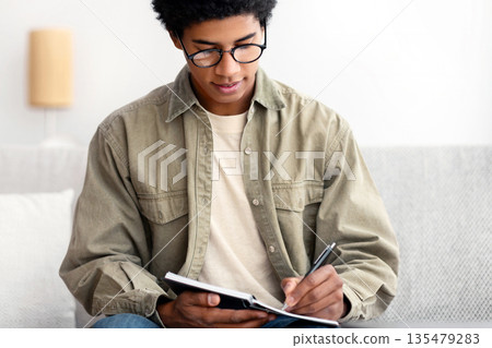 Busy African American teenage guy working on educational project at home. Black male student studying remotely, writing home assignment in notebook, taking notes for college test Busy African American teenage guy working on educational project at home. Black male student studying remotely, writing home assignment in notebook, taking notes for college test 135479283