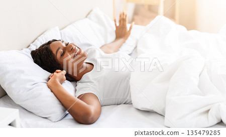 Happy african american young woman waking up in her comfortable bed at home, stretching and smiling, copy space. Cheerful black lady starting her morning with smile, feeling good and lifeful 135479285