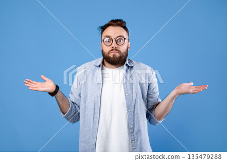 Who knows. Confused bearded man in glasses shrugging shoulders on blue studio background. Unsure millennial guy expressing uncertainty, feeling clueless, making hard choice 135479288