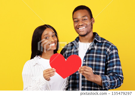 Valentines day, anniversary and modern relationship. Smiling millennial african american guy and lady hold red heart at self-isolation, isolated on yellow background, studio shot, copy space Valentines day, anniversary and modern relationship. Smiling millennial african american guy and lady hold red heart at self-isolation, isolated on yellow background, studio shot, copy space 135479297