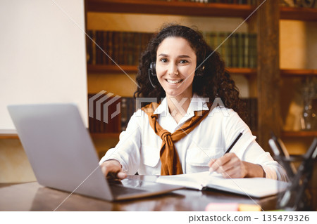 E-learning And Distant Education During Quarantine. Positive young female student in headset studying online, using laptop, writing and taking notes in her notebook, looking and posing at camera E-learning And Distant Education During Quarantine. Positive young female student in headset studying online, using laptop, writing and taking notes in her notebook, looking and posing at camera 135479326