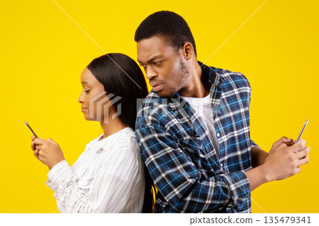 Jealous man checks female phone. Strict millennial african american guy look, wife in casual, texting on smartphone and has romantic online relationship, isolated on yellow background, studio shot Jealous man checks female phone. Strict millennial african american guy look, wife in casual, texting on smartphone and has romantic online relationship, isolated on yellow background, studio shot 135479341