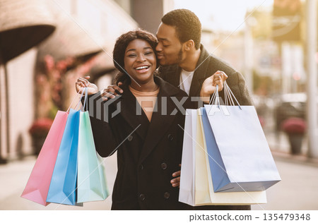 Love and shopping together during big sale at Black Friday in november. Happy millennial african american man kisses lady with many colored packages near modern city shop before holidays, copy space 135479348