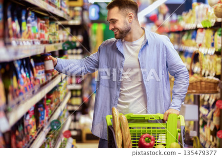 Everyday Shopping Concept. Smiling Young Man Pushing Trolley Cart Between Aisles In Grocery Store. Positive Guy Buying Food, Taking Products From Shelf. Consumerism And Lifestyle Everyday Shopping Concept. Smiling Young Man Pushing Trolley Cart Between Aisles In Grocery Store. Positive Guy Buying Food, Taking Products From Shelf. Consumerism And Lifestyle 135479377