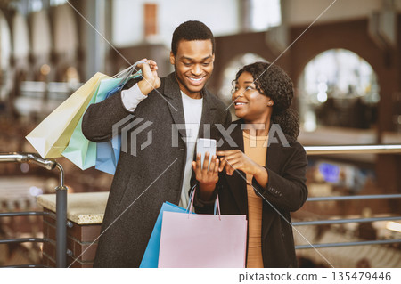 Black Friday and mobile app for online shopping and cash back. Cheerful african american young woman with colorful packages shows to man smartphone with message about sale and discount at urban mall Black Friday and mobile app for online shopping and cash back. Cheerful african american young woman with colorful packages shows to man smartphone with message about sale and discount at urban mall 135479446