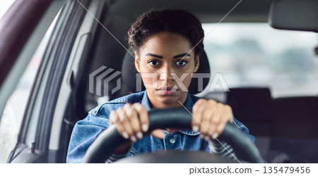 Facial expression, negativity, stress and traffic jams. Serious upset young african american lady sitting in car behind steering wheel and looking ahead tensely in window driving car, free space 135479456