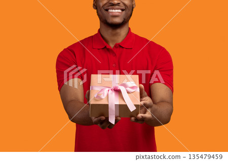 This Present Is For You. Closeup cropped view of unrecognizable smiling black bearded man holding wrapped gift box with pink ribbon, giving and showing to camera, isolated on orange studio background 135479459
