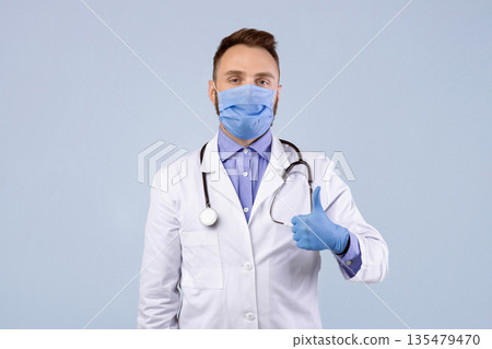 Medical doctor in lab coat, protective mask and rubber gloves showing thumb up gesture over grey studio background. General practitioner wearing protection during covid-19 epidemic Medical doctor in lab coat, protective mask and rubber gloves showing thumb up gesture over grey studio background. General practitioner wearing protection during covid-19 epidemic 135479470