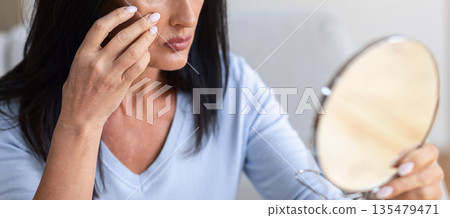 Skin Aging. Closeup portrait of sad mature brunette woman holding mirror and looking at her face in reflection, applying moisturizing cream, touching wrinkles, preparing facial skin for makeup 135479471