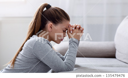 Side view of desperate young woman praying for help or consolation on sofa at home, copy space. Religious millennial lady worshiping God, asking for support in her grief or loss 135479476