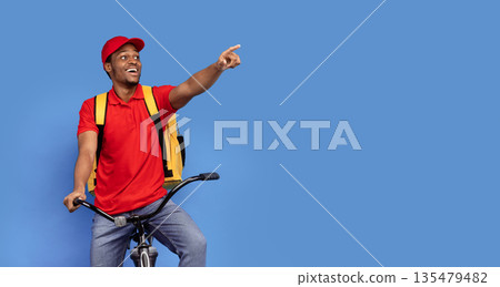 Look Here. Emotional surprised black delivery man in red uniform riding bicycle, wearing thermo backpack bag and pointing away at copy space isolated on blue studio background, banner, panorama 135479482