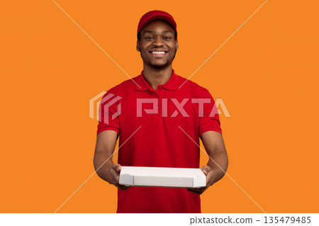 Fast Food Delivery. Portrait of smiling african american male courier wearing red cap and uniform holding pizza box from restaurant and giving it to camera, isolated on orange yellow background Fast Food Delivery. Portrait of smiling african american male courier wearing red cap and uniform holding pizza box from restaurant and giving it to camera, isolated on orange yellow background 135479485