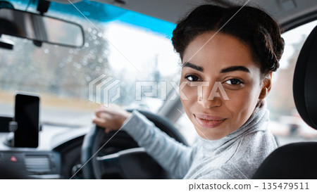 Female driver talking to passenger in back seat or auto blogger. Cheerful attractive young african american woman holding steering wheel, turns and looks over her shoulder in camera, empty space 135479511
