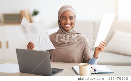 Positive black Muslim woman in hijab working from home, holding documents near laptop computer. Portrait of attractive African American lady in headcarf doing paperwork, indoors Positive black Muslim woman in hijab working from home, holding documents near laptop computer. Portrait of attractive African American lady in headcarf doing paperwork, indoors 135479530