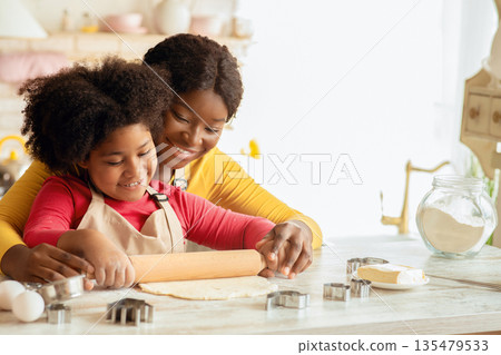 Happy black mom and her daughter rolling up dough together on table in kitchen, having fun while preparring homemade pastry, loving african american family enjoying spending time together at home Happy black mom and her daughter rolling up dough together on table in kitchen, having fun while preparring homemade pastry, loving african american family enjoying spending time together at home 135479533