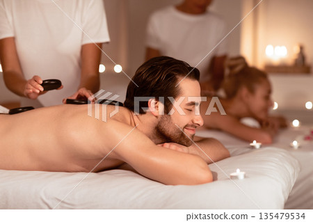 Spouses Relaxing Receiving Exotic Hot Stones Massage Lying In Luxury Spa Resort. Wellness And Relaxation During Romantic Honeymoon Vacation. Low Light, Selective Focus Spouses Relaxing Receiving Exotic Hot Stones Massage Lying In Luxury Spa Resort. Wellness And Relaxation During Romantic Honeymoon Vacation. Low Light, Selective Focus 135479534