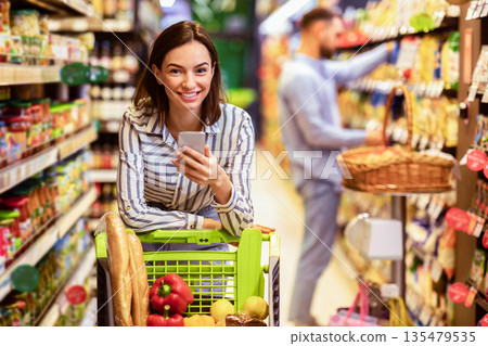 Portrait of smiling young casual woman leaning on shopping trolley cart indoors, using her mobile phone, posing and looking at camera. Satisfied female customer buying products in local grocery store 135479535