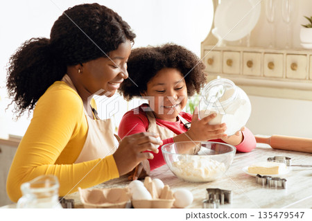 Cute Little Black Girl Baking In Kitchen With Her Mom, Adding Flour To Bowl While Preparing Dough For Cookies, Happy African American Mother And Daughter Having Fun Together At Home, Free Space 135479547