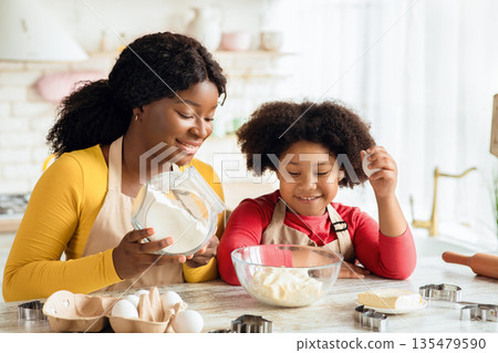 Cheerful black mother and little daughter preparing dough in kitchen together, adding ingredients to bowl, enjoying making homemade pastry, cute girl and her mom wearing aprons having fun at home 135479590