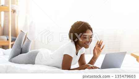 Cute black lady laying on bed, wearing pajamas, having video call on computer from home, panorama. Cheerful african american young woman waving at laptop screen and smiling, copy space 135479603