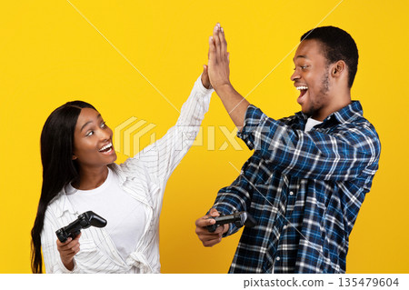 Victory together, love, friendship and modern entertainment. Joyful excited zoomers african american male and female give high five with gamepads in hands, isolated on yellow background, studio shot 135479604