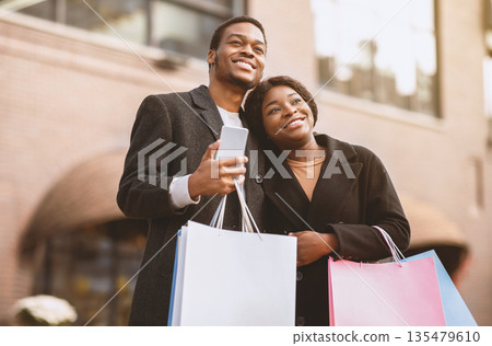 Great shopping on black friday and advertising of sale. Happy african american man and lady in coats with colored bags hug and look at something amazing, male holding smartphone near shop, copy space Great shopping on black friday and advertising of sale. Happy african american man and lady in coats with colored bags hug and look at something amazing, male holding smartphone near shop, copy space 135479610