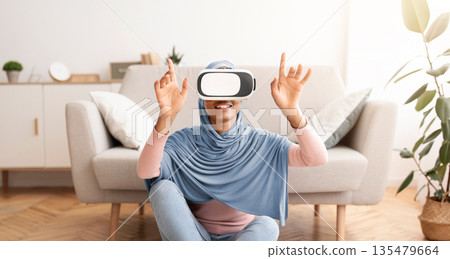 Weekend pastimes. Young Muslim woman in hijab interacting with virtual reality, wearing VR headset at home. African American lady in traditional headscarf playing video game in cyberspace Weekend pastimes. Young Muslim woman in hijab interacting with virtual reality, wearing VR headset at home. African American lady in traditional headscarf playing video game in cyberspace 135479664