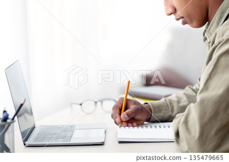 Remote education concept. Side view of black teen guy taking notes in copybook during web conference at home. African American adolescent taking online lesson or listening to lecture on internet Remote education concept. Side view of black teen guy taking notes in copybook during web conference at home. African American adolescent taking online lesson or listening to lecture on internet 135479665