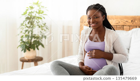Maternity Concept. Portrait Of Happy Expectant Black Lady Posing On Bed At Home, Embracing Her Pregnant Belly And Smiling At Camera, Positive Awaiting Woman Enjoying Her Healthy Pregnancy, Free Space 135479669
