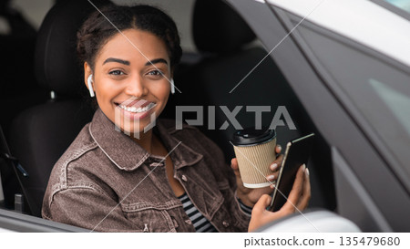 Modern driver on break with gadgets and coffee. Happy young african american lady sits in car with cup of hot drink takeaway, in wireless headphones with smartphone and looking at camera, free space Modern driver on break with gadgets and coffee. Happy young african american lady sits in car with cup of hot drink takeaway, in wireless headphones with smartphone and looking at camera, free space 135479680