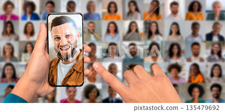 A user interacts with a smartphone, scanning their face for identification. Behind them, a collage features various diverse portraits illustrating modern facial recognition technology. A user interacts with a smartphone, scanning their face for identification. Behind them, a collage features various diverse portraits illustrating modern facial recognition technology. 135479692