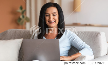 Domestic Comfort. Portrait Of Positive Mature Woman Using And Working On Laptop At Home, Enjoying Free Time, Smiling Lady Sitting On The Couch With Computer On Her Lap, Watching Video Domestic Comfort. Portrait Of Positive Mature Woman Using And Working On Laptop At Home, Enjoying Free Time, Smiling Lady Sitting On The Couch With Computer On Her Lap, Watching Video 135479693