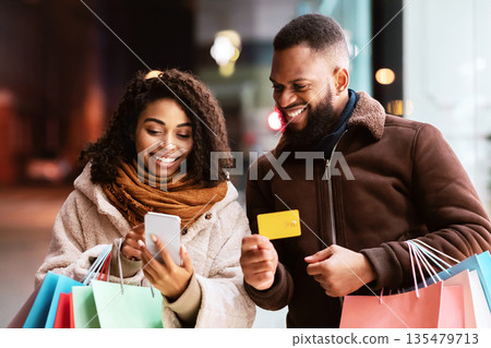 Ordering Online. Smiling black casual couple using cellphone, man showing debit credit card, family holding colorful shopping bags, walking outdoors in the evening, making purchases on internet Ordering Online. Smiling black casual couple using cellphone, man showing debit credit card, family holding colorful shopping bags, walking outdoors in the evening, making purchases on internet 135479713
