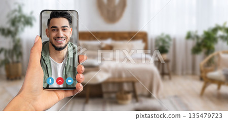 A person holds a smartphone displaying a smiling individual during a video call from their cozy home interior. The background features a warm, inviting bedroom environment, emphasizing connection. A person holds a smartphone displaying a smiling individual during a video call from their cozy home interior. The background features a warm, inviting bedroom environment, emphasizing connection. 135479723