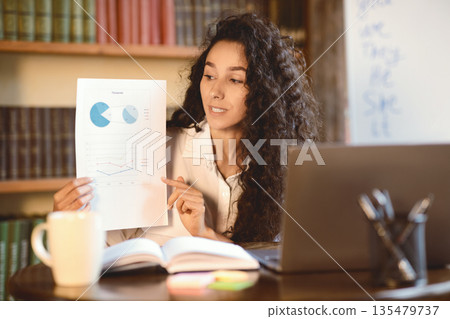 Portrait of female financial analyst having video call on laptop, giving virtual presentation. Woman showing statistics, graphs and charts to camera, holding document paper, explaining report Portrait of female financial analyst having video call on laptop, giving virtual presentation. Woman showing statistics, graphs and charts to camera, holding document paper, explaining report 135479737