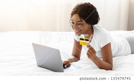 Online shopping from home, e-commerce concept. Happy young african american woman lying on bed at home and buying clothes or products online, paying with credit card via laptop, copy space 135479745