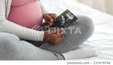 Fetal Development. Pregnant african american woman looking at ultrasound scan of baby at home, holding sonography image in hands, sitting on bed, enjoying healthy pregnancy, cropped image, closeup Fetal Development. Pregnant african american woman looking at ultrasound scan of baby at home, holding sonography image in hands, sitting on bed, enjoying healthy pregnancy, cropped image, closeup 135479746