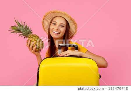 Pretty young woman with pineaaple and yellow luggage posing over pink studio background, copy space. Happy lady traveller in summer hat going to warm seaside. Destinations to travel concept Pretty young woman with pineaaple and yellow luggage posing over pink studio background, copy space. Happy lady traveller in summer hat going to warm seaside. Destinations to travel concept 135479748