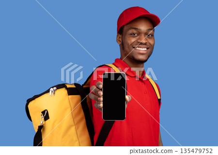 Application For Online Order. Smiling black deliveryman showing mobile phone with blank screen for mockup, wearing red cap, uniform and thermo backpack bag, isolated on blue background Application For Online Order. Smiling black deliveryman showing mobile phone with blank screen for mockup, wearing red cap, uniform and thermo backpack bag, isolated on blue background 135479752