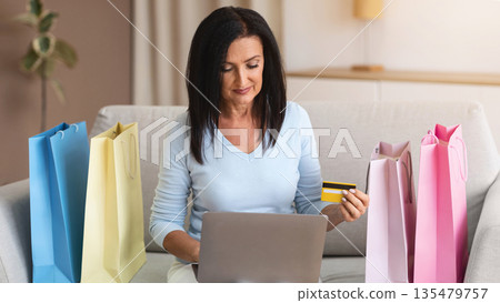 Online Transaction Concept. Confident mature lady holding debit credit card and using laptop, ordering clothes and purchases via internet, sitting on sofa with colorful shopping bags in living room Online Transaction Concept. Confident mature lady holding debit credit card and using laptop, ordering clothes and purchases via internet, sitting on sofa with colorful shopping bags in living room 135479757