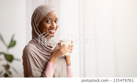 Black Muslim woman in traditional hijab drinking hot aromatic coffee near window at home, having peaceful weekend. Portrait of beautiful young lady in Arabic headscarf having warming beverage indoors Black Muslim woman in traditional hijab drinking hot aromatic coffee near window at home, having peaceful weekend. Portrait of beautiful young lady in Arabic headscarf having warming beverage indoors 135479761