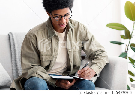Distance education. African American teen university student studying from home at times of lockdown. Black teenager taking notes for his test, making home assignment or preparing for online lesson Distance education. African American teen university student studying from home at times of lockdown. Black teenager taking notes for his test, making home assignment or preparing for online lesson 135479766
