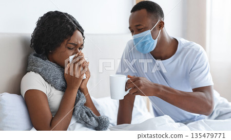 Black man in protective face mask taking care of his sick wife, giving hot drink, bedroom interior, empty space. African american woman with scarf around her neck having coronavirus, sneezing her noze Black man in protective face mask taking care of his sick wife, giving hot drink, bedroom interior, empty space. African american woman with scarf around her neck having coronavirus, sneezing her noze 135479771
