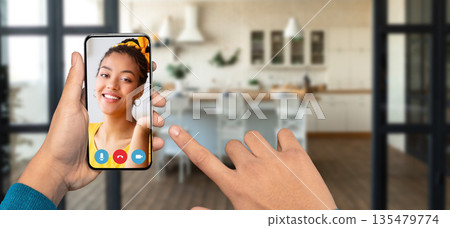 A person holds a smartphone in their hand, has video call from their home. The cozy background features a modern kitchen. A person holds a smartphone in their hand, has video call from their home. The cozy background features a modern kitchen. 135479774