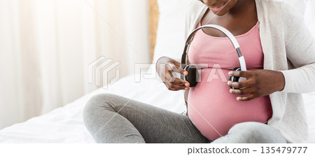 Black Pregnant Woman Holding Headphones Near Belly, Playing Melody To Baby In Womb, Enjoying Listening Classical Music During Pregnancy, Sitting On Bed At Home, Closeup Shot With Free Space Black Pregnant Woman Holding Headphones Near Belly, Playing Melody To Baby In Womb, Enjoying Listening Classical Music During Pregnancy, Sitting On Bed At Home, Closeup Shot With Free Space 135479777